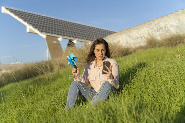 Retrato de una joven sonriente sentada en un prado con un molinete mirando el teléfono móvil - foto de stock
