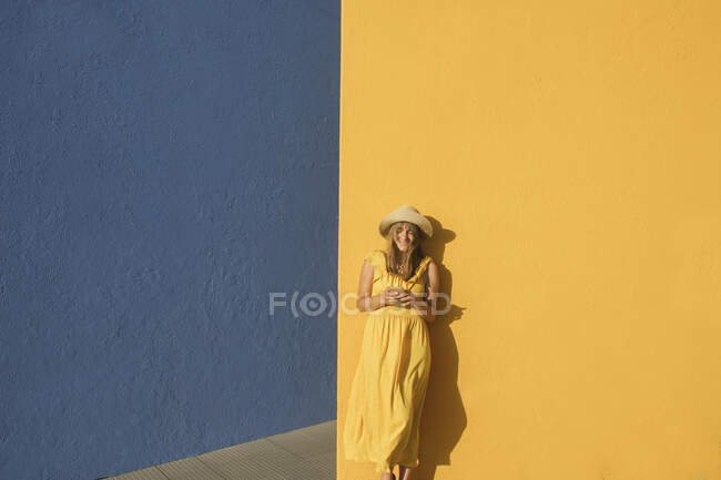 Mujer en un vestido amarillo usando smartphone frente a paredes amarillas y azules - foto de stock