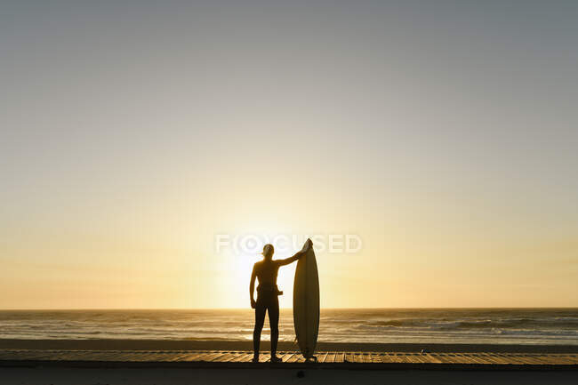 Surfista de pie con la tabla de surf frente a la puesta de sol en la playa - foto de stock
