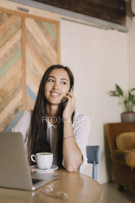 Молода бізнес-леді з ноутбуком розмовляє по телефону за столом в кафе — стокове фото