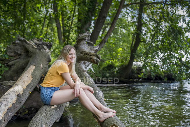Mujer sonriente sentada en el tronco de un árbol a orillas del lago - foto de stock