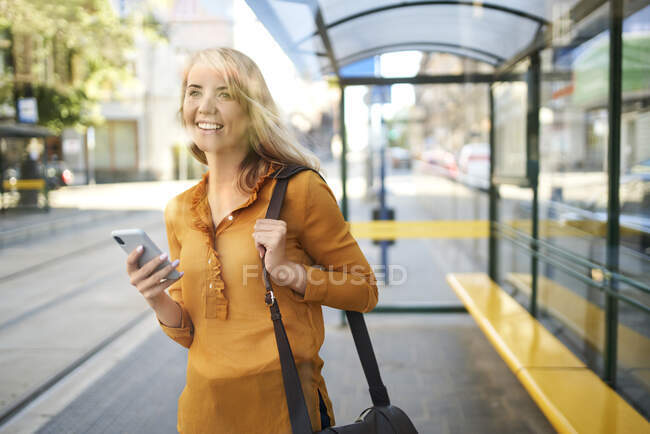 Sorrindo jovem com smartphone esperando o bonde na parada de bonde — Fotografia de Stock