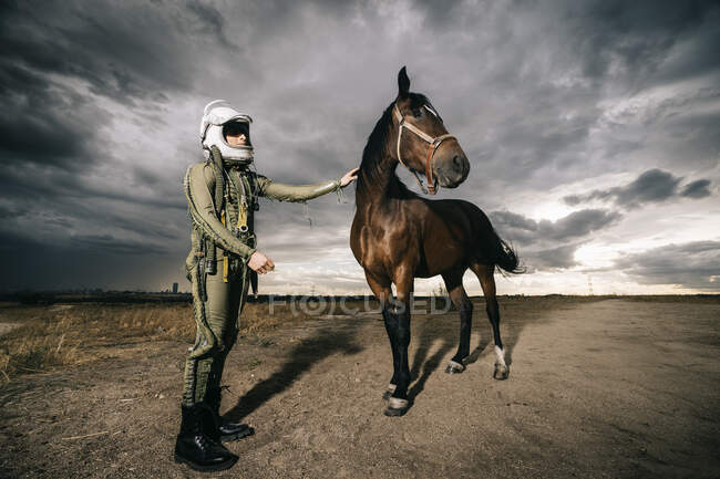 Hombre vestido como un astronauta con un caballo en un prado con nubes dramáticas en el fondo - foto de stock