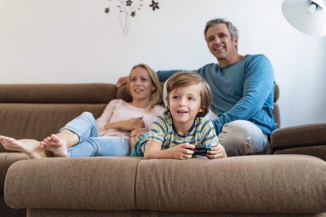 Menino deitado no sofá em casa jogando videogame com os pais assistindo — Fotografia de Stock