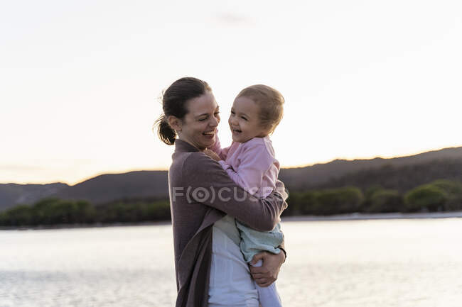 Happy mother carrying her daughter on a jetty at sunset — Stock Photo