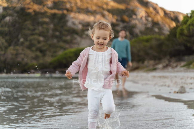 Chica feliz corriendo en la orilla del mar con el padre en el fondo - foto de stock