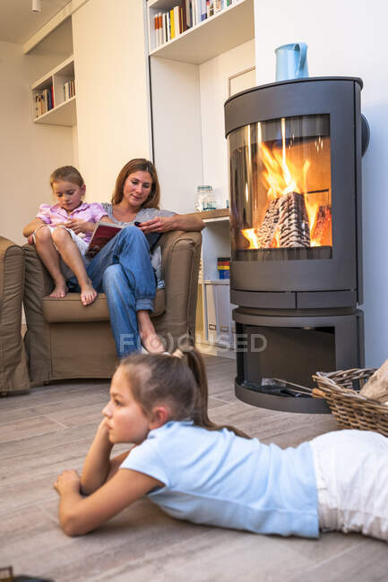 Madre con hijas relajándose y leyendo en la sala de estar en la chimenea - foto de stock