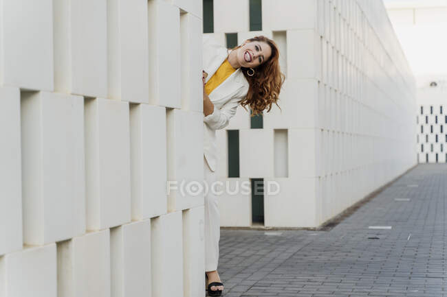 Femme d'affaires en costume de pantalon blanc, debout derrière le mur, sortant la langue — Photo de stock