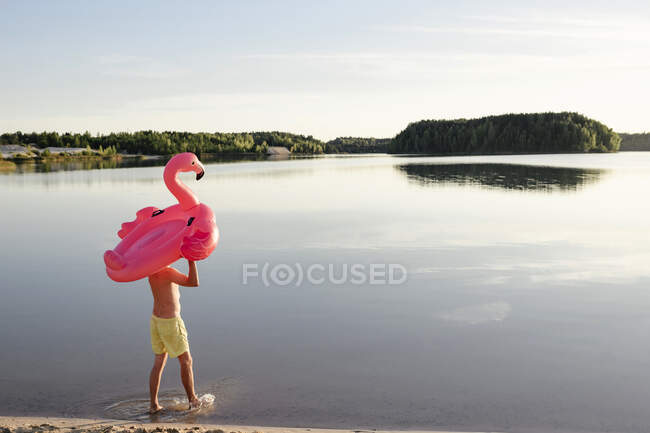 Young man with flamingo pool float walking at lakeshore — Stock Photo