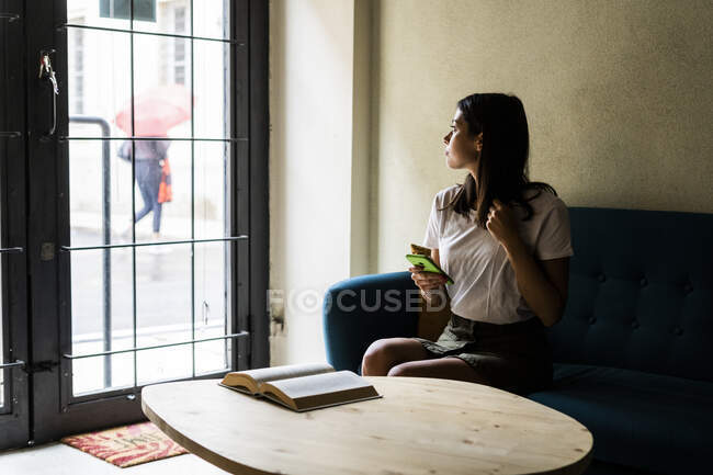 Молода жінка з мобільним телефоном сидить на дивані і дивиться у вікно. — стокове фото