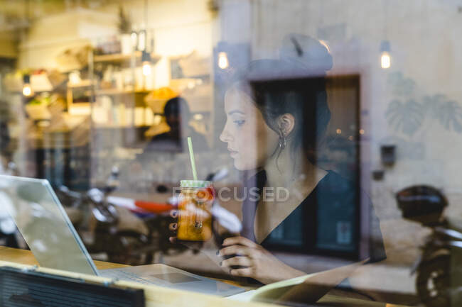 Young woman with smoothie and laptop behind windowpane in a cafe — Stock Photo