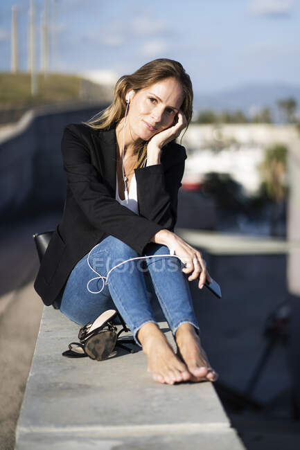 Porträt einer selbstbewussten Geschäftsfrau, die mit Smartphone und Kopfhörer an der Wand sitzt — Stockfoto