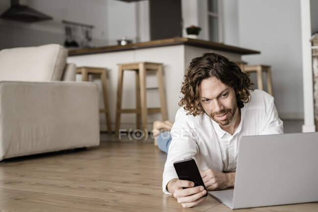 Man lying on the floor at home using cell phone and laptop — Stock Photo