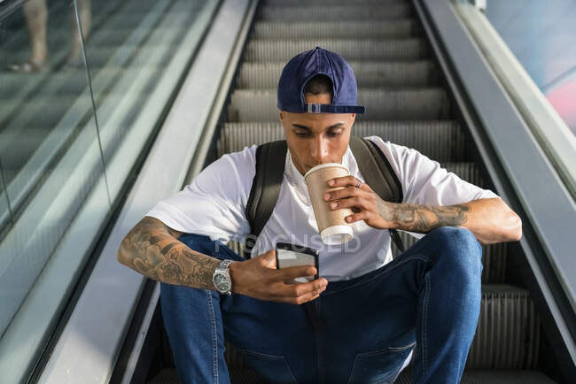 Молодий чоловік з рюкзаком сидить на ескалаторі і дивиться на мобільний телефон, коли п'є каву. — стокове фото