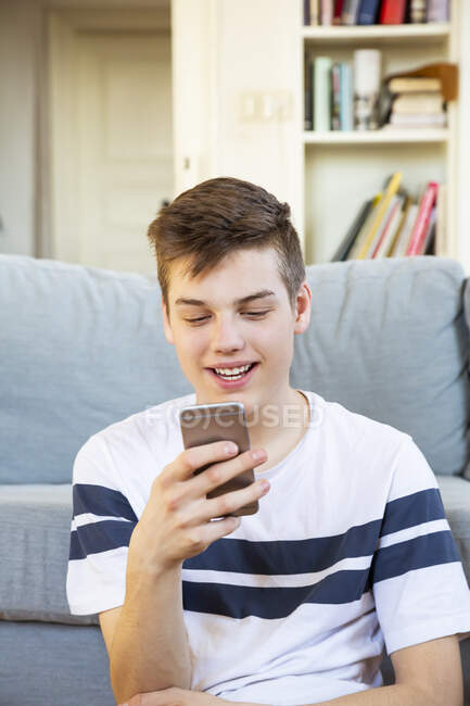 Портрет усміхненого підлітка, який сидить навпроти дивана за допомогою мобільного телефону. — стокове фото
