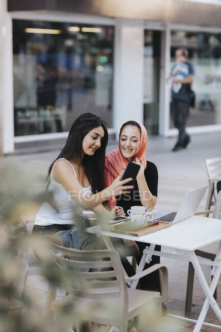 Two friends sitting together at a pavement cafe using cell phone — Stock Photo