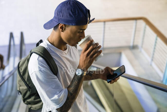 Татуювання молодого чоловіка з рюкзаком стоїть на ескалаторі, дивлячись на мобільний телефон, і п'є каву, щоб піти. — стокове фото