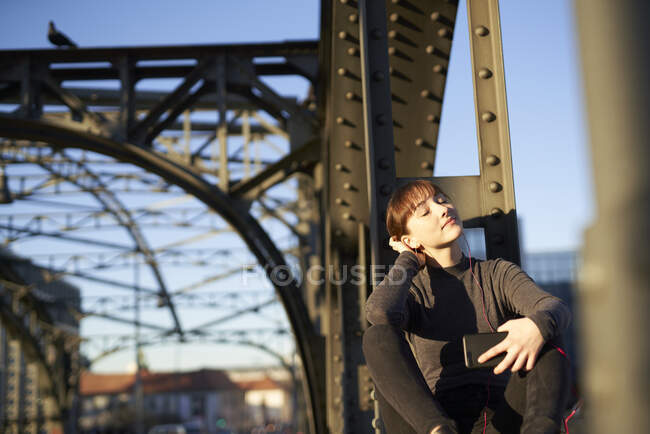 Ritratto di giovane donna rilassata seduta sulla trave d'acciaio di un ponte che ascolta musica con smartphone — Foto stock