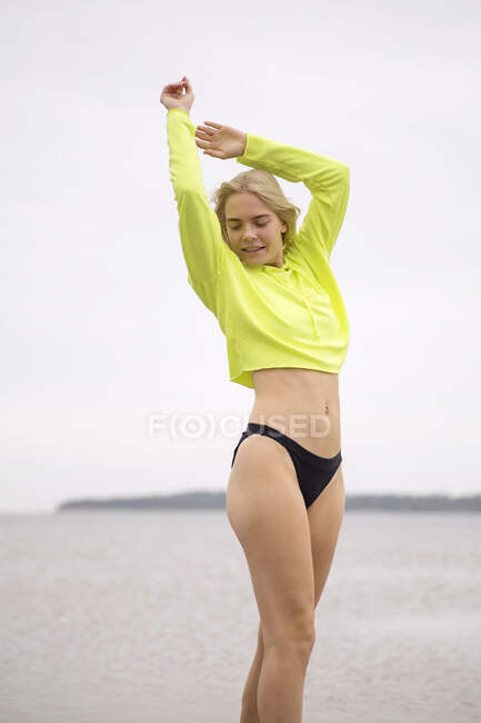 Ritratto di giovane donna bionda in posa sulla spiaggia con felpa gialla al neon e pantaloni bikini neri — Foto stock