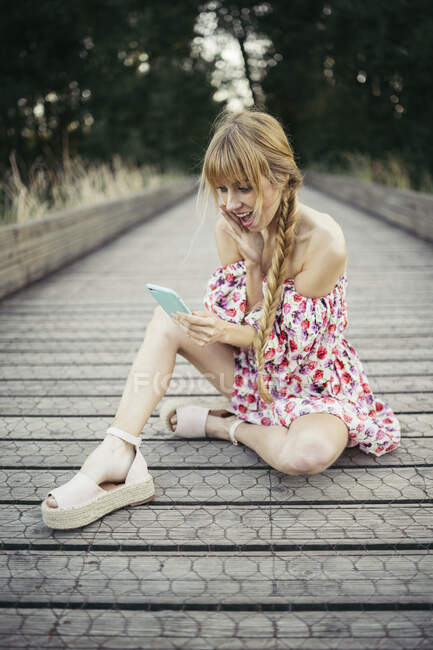 Porträt einer überraschten jungen Frau, die auf der Uferpromenade sitzt und aufs Smartphone schaut — Stockfoto
