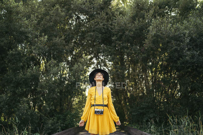 Young woman wearing a black hat and yellow dress with an analog camera on wooden boardwalk — Stock Photo