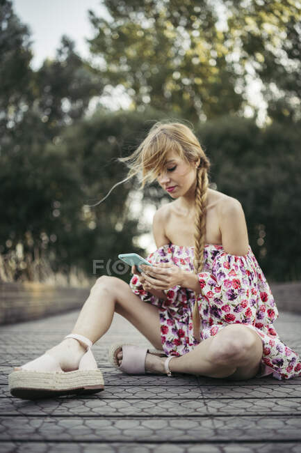Retrato de mujer joven con vestido de verano con diseño floral sentado en el paseo marítimo usando el teléfono celular - foto de stock