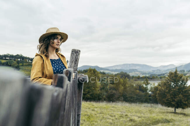Jovem de cabelos encaracolados mulher vestindo um chapéu, casaco amarelo e camiseta azul olhando a paisagem da montanha — Fotografia de Stock