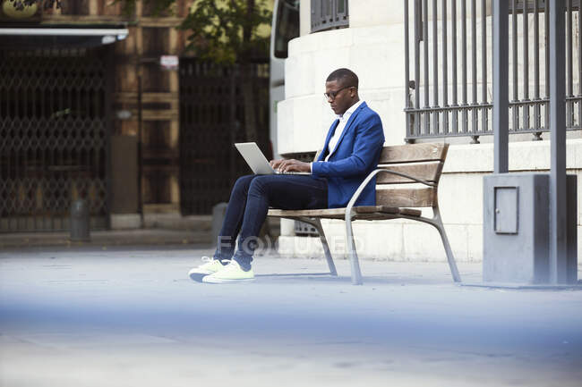 Young businessman wearing blue suit jacket sitting on bench and using laptop - foto de stock