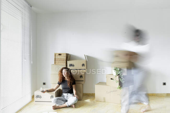 Молодая женщина сидит в новом доме с бокалом красного вина, в то время как друг быстро движется с картонной коробкой — стоковое фото