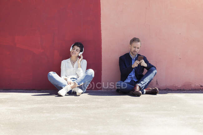 Couple leaning on a wall, man using smartphone, woman with white headphones — Stock Photo
