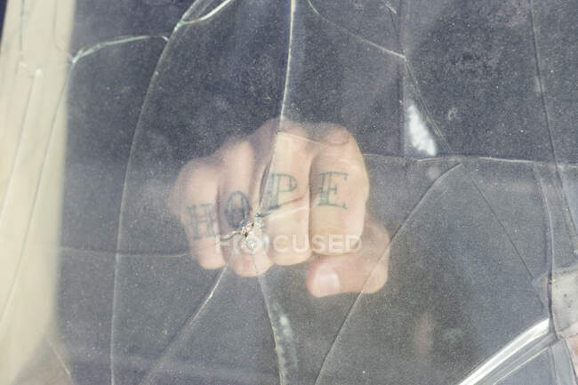 Fist of a young man with HOPE tattoo behind broken windscreen on a scrapyard — Stock Photo
