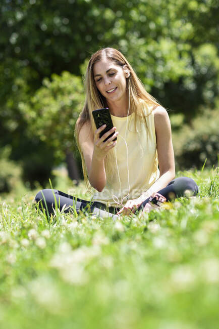 Mujer usando su smartphone, descansando en la hierba de un parque después de hacer ejercicio - foto de stock