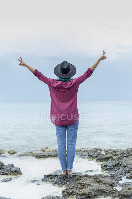 Back view of young woman with dyed blue hair standing on rock in front of the sea showing victory signs, Spain — Stock Photo