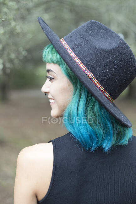 Lächelnde junge Frau mit gefärbten blauen und grünen Haaren mit Hut an einem regnerischen Tag — Stockfoto