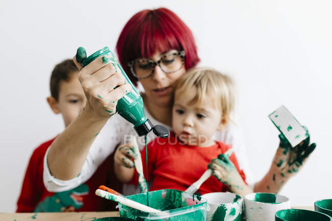 Madre con sus hijos haciendo manualidades en casa y pintando con verde - foto de stock