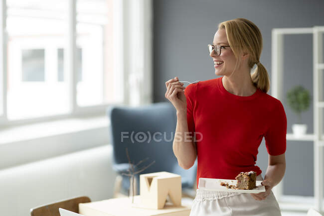 Glückliche junge Frau isst im Büro ein Stück Kuchen — Stockfoto