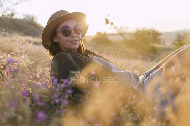 Young woman wearing sunglasses and sunhat, sitting on meadow at sunset — Stock Photo