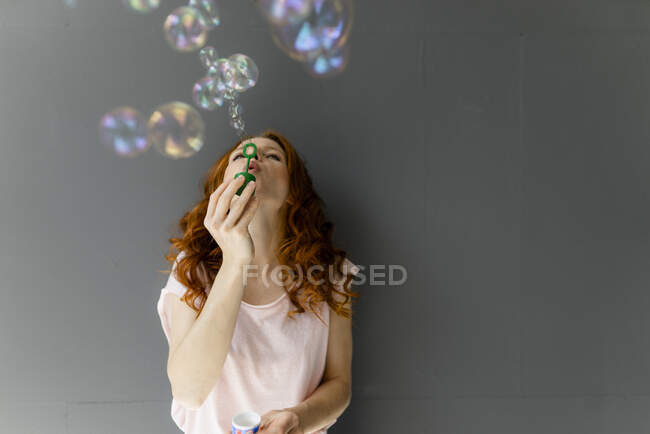Donna dai capelli rossi appoggiata al muro grigio che soffia bolle di sapone — Foto stock