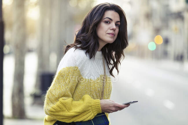 Brunette woman looking to cross the street while holding her smartphone in the city — Stock Photo