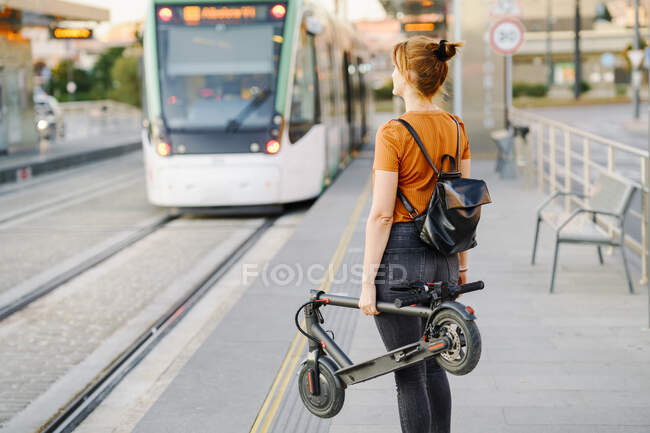 Visão traseira da mulher com mochila e scooter elétrico dobrado esperando na parada de bonde — Fotografia de Stock