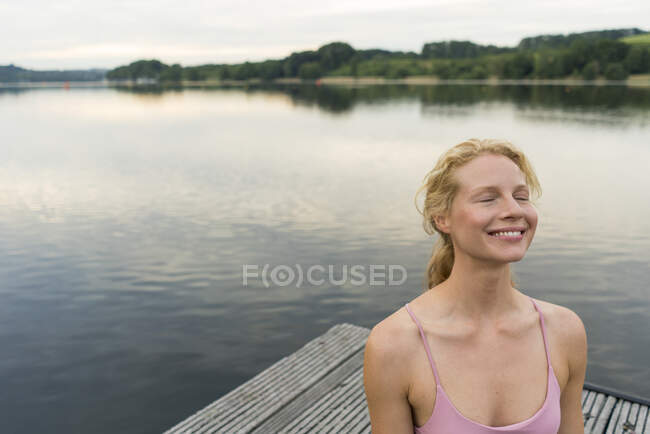 Sorridente giovane donna con gli occhi chiusi su un molo a un lago — Foto stock
