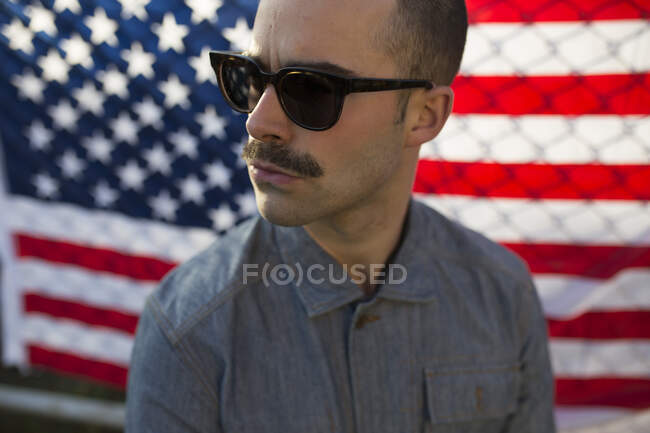 Giovane uomo con occhiali da sole davanti alla bandiera statunitense sulla recinzione — Foto stock
