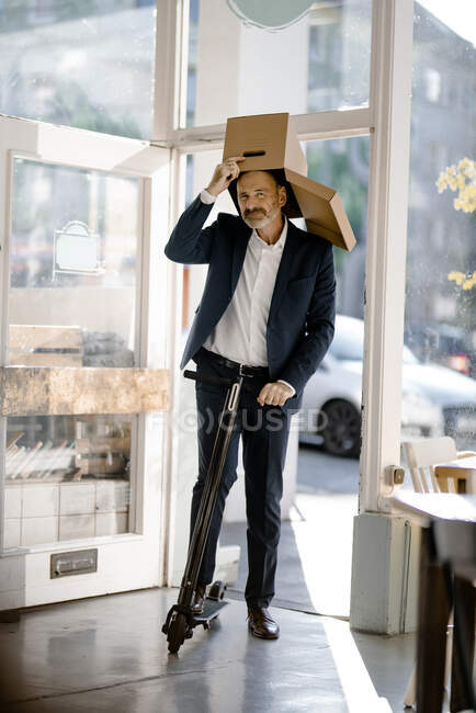 Бизнесмен с картонной коробкой на голове, ездит на электронном скутере в кофейне — стоковое фото