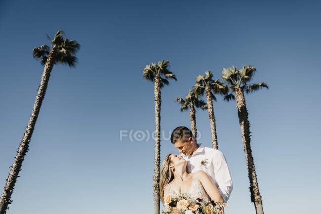 Happy bride and groom hugging at palm trees under blue sky — Stock Photo