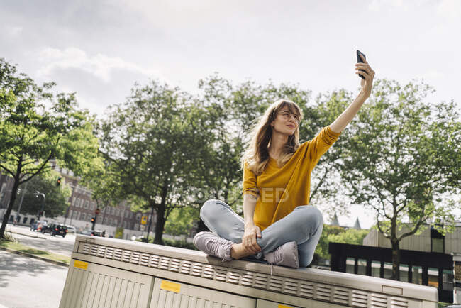 Jovem mulher sentada em uma caixa na cidade tirando uma selfie — Fotografia de Stock
