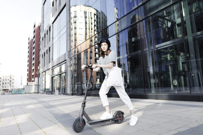 Жінка з електронним скутером і шолом, сучасні офісні будівлі на задньому плані — стокове фото