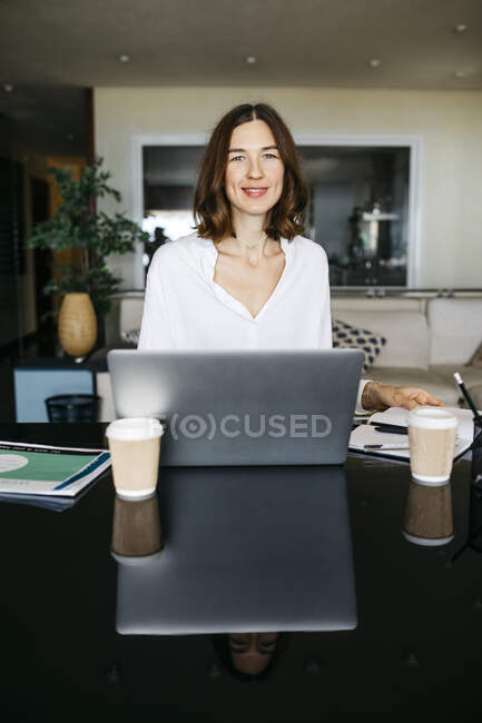Retrato de la mujer sonriente trabajando en la mesa en casa con el ordenador portátil - foto de stock
