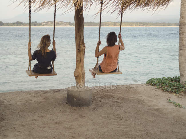 Vista trasera de dos mujeres sentadas en columpios frente al mar, Islas Gili, Bali - foto de stock