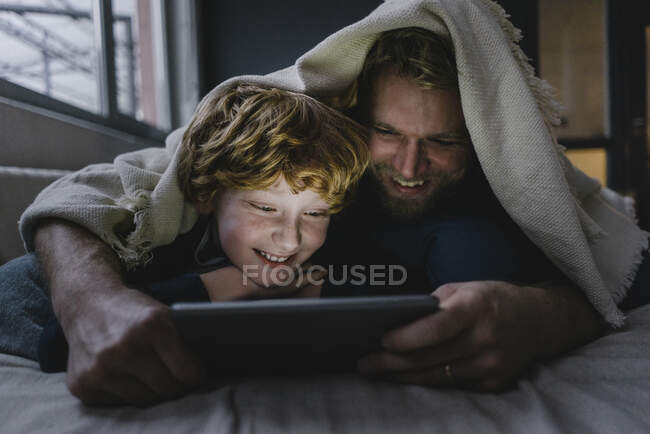 Padre e figlio sdraiati insieme sotto una coperta guardando tablet digitale — Foto stock