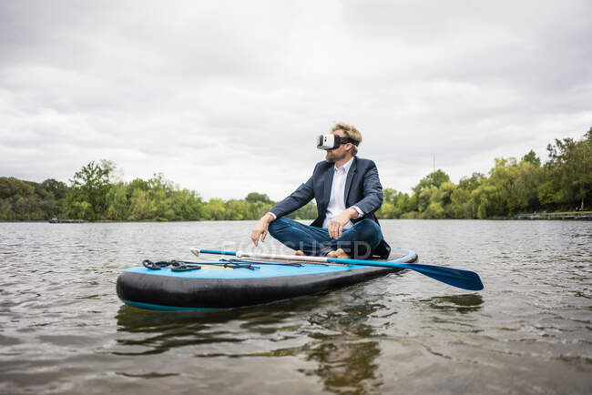 Бизнесмен в очках VR сидит на доске SUP на озере — стоковое фото